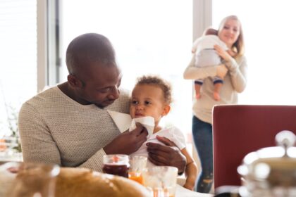 Dad holding small baby on his lap while mum looks on from behind him holding another baby